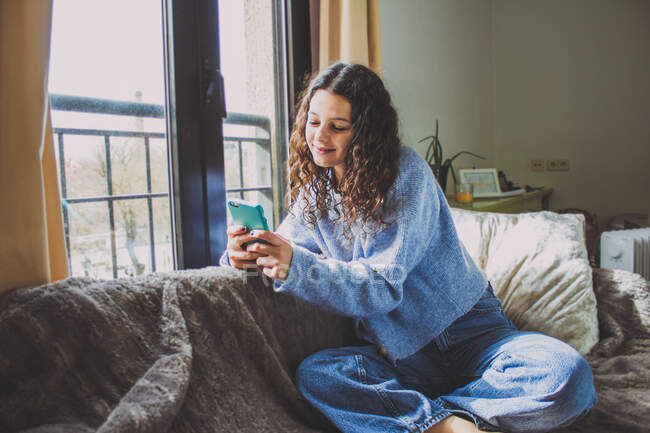 Молода жінка дивиться на мобільний на дивані біля вікна — стокове фото