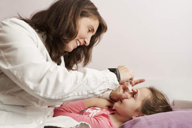 Doctora sonriendo y dando gotas para los ojos a una niña en su cama. Concepto médico casero - foto de stock