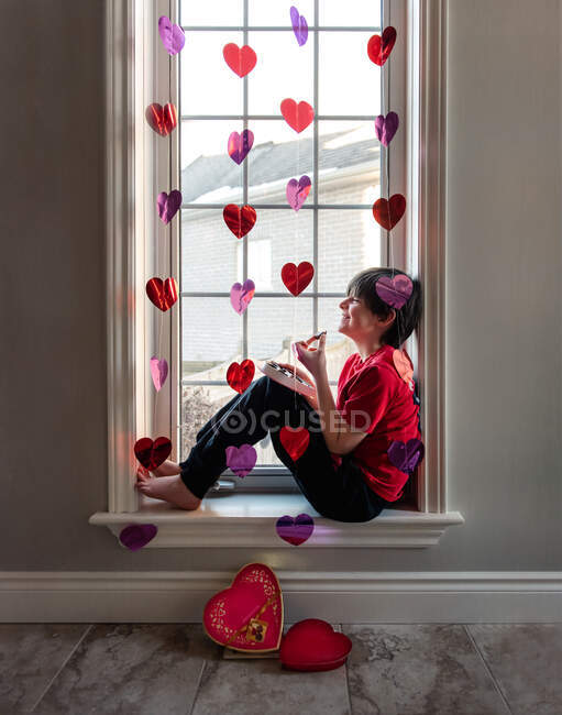 Menino comendo chocolates na janela decorada com corações para Valentines. — Fotografia de Stock