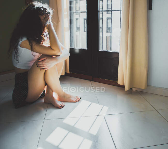 Молодая женщина, сидящая обеспокоенная на полу и выглядывающая в окно — стоковое фото