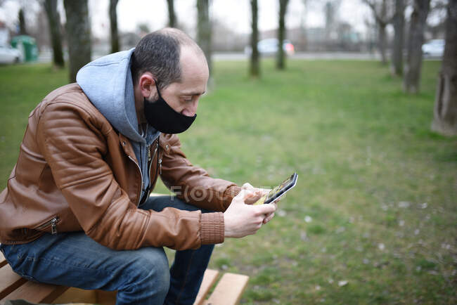 Caucásico hombre usando una máscara para protegerse de covid19 mira a su teléfono celular en un banco de madera parque, espacio abierto. - foto de stock