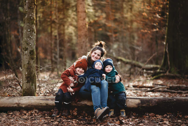 Mamá e hijos sonriendo felizmente sentados en el bosque en el otoño - foto de stock