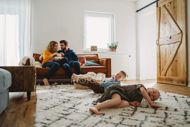 Siblings having fun in living room parents sit on sofa with baby — Stock Photo