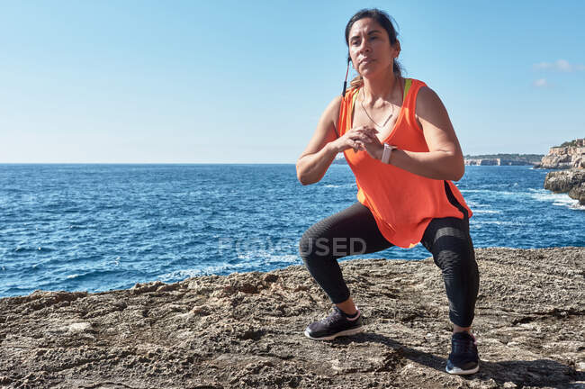 Latin woman, middle-aged, with orange top, black leggings, training, doing physical exercises, squats, burning calories, keeping fit, outdoors by the sea, blue sky, day, sunny winter, bluetoo — Stock Photo