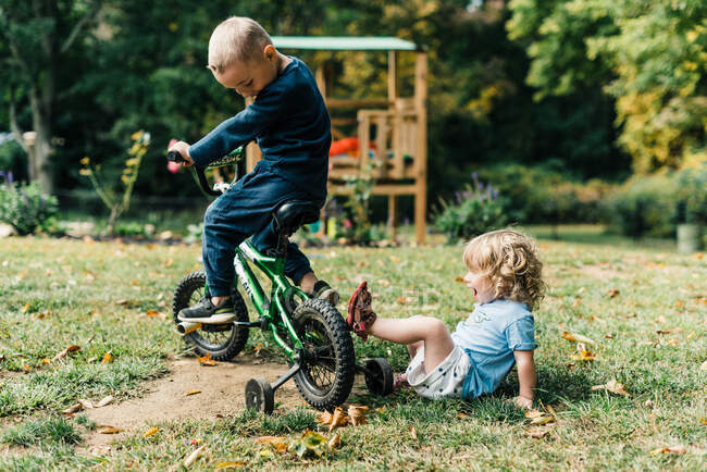 Dos niños pequeños, hermanos, jugando en el patio trasero con una bicicleta en la tierra - foto de stock