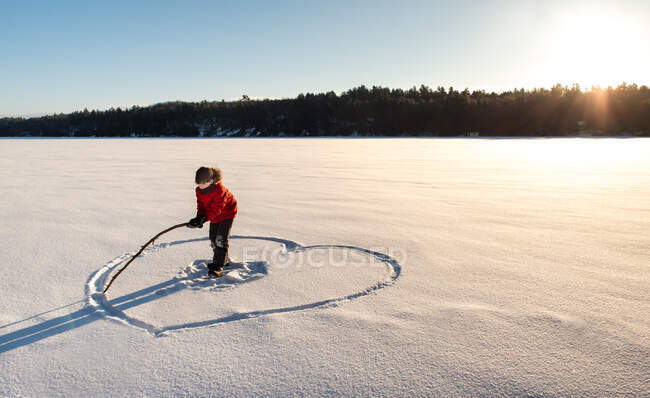 Niño dibujando una forma de corazón en un campo cubierto de nieve al sol de la mañana. - foto de stock