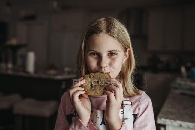 Tween girl eating a large chocolate chip cookie in kitchen with apron — Stock Photo