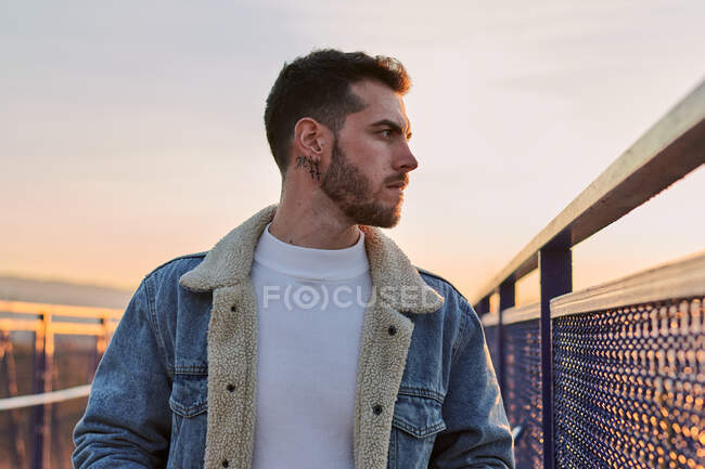 Homem bonito em uma jaqueta posa em uma ponte ao pôr do sol — Fotografia de Stock