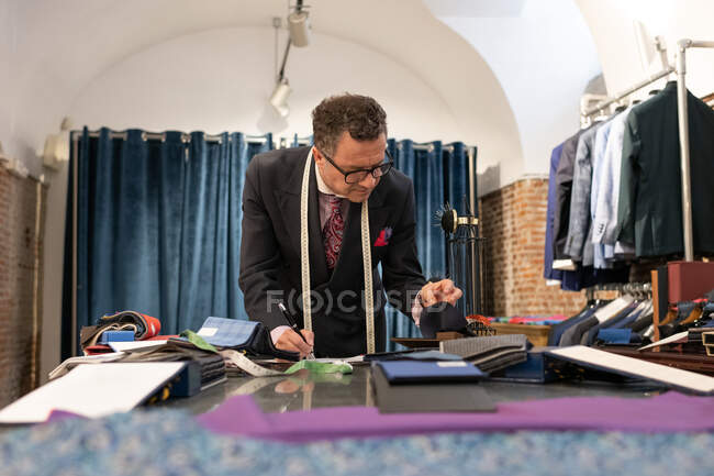 Старший человек в элегантном костюме подбирает одежду и пишет во время работы в портной студии — стоковое фото