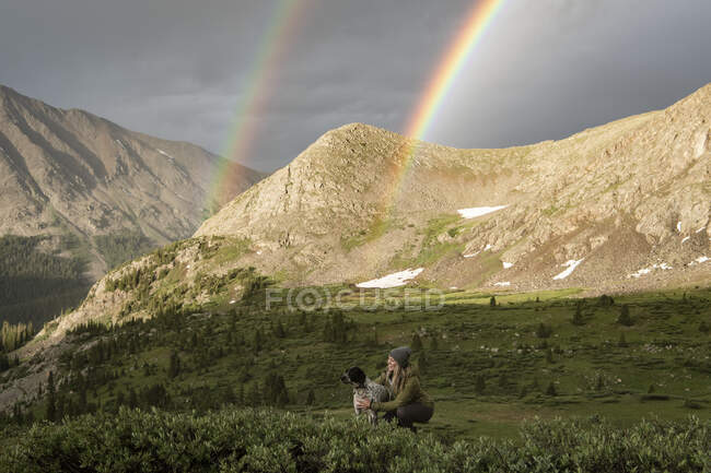 Woman stroking dog while double rainbow and mountains in background — Stock Photo