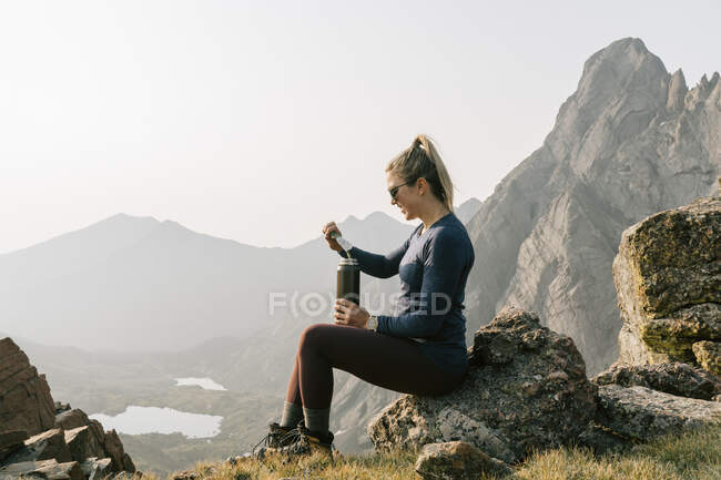 Vue latérale de la femme boire de l'énergie tout en étant assis sur la montagne contre le ciel clair — Photo de stock