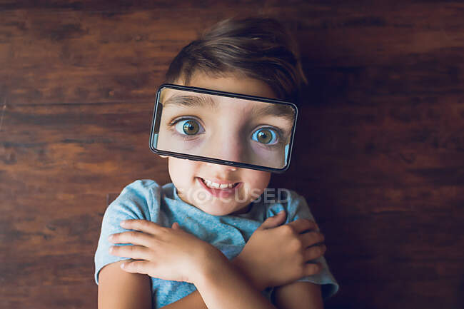 Niño tendido en el suelo con una foto de teléfono de sus ojos en la parte superior de la cara - foto de stock