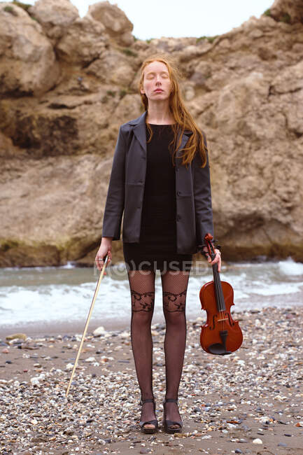 Violinista pelirroja posando con su violín en la playa en un día nublado - foto de stock