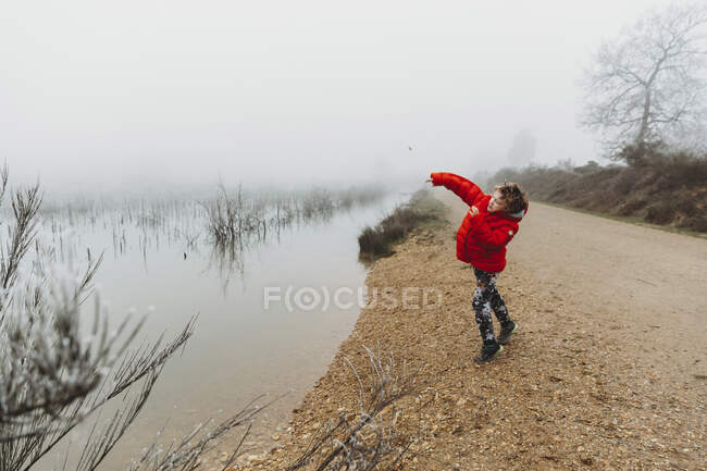 Niño con abrigo rojo arrojando piedra al lago contra el cielo nublado de niebla - foto de stock