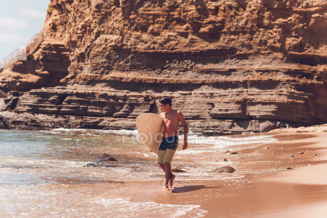 Niño caminando en la playa llevando su tabla de surf - acantilados en la parte posterior. - foto de stock