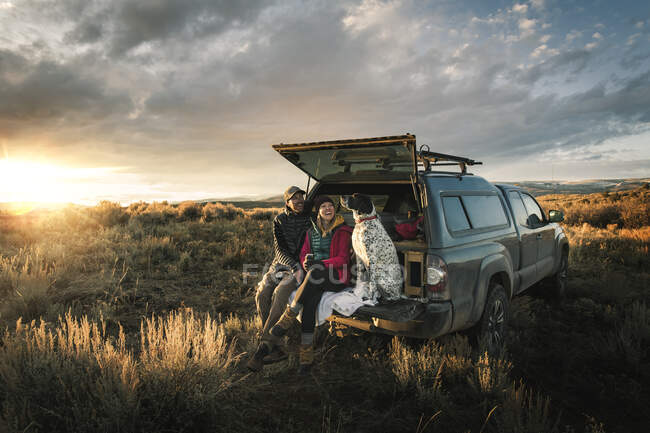 Alegre pareja sentada con perro en el maletero del vehículo todoterreno en el campo durante las vacaciones - foto de stock