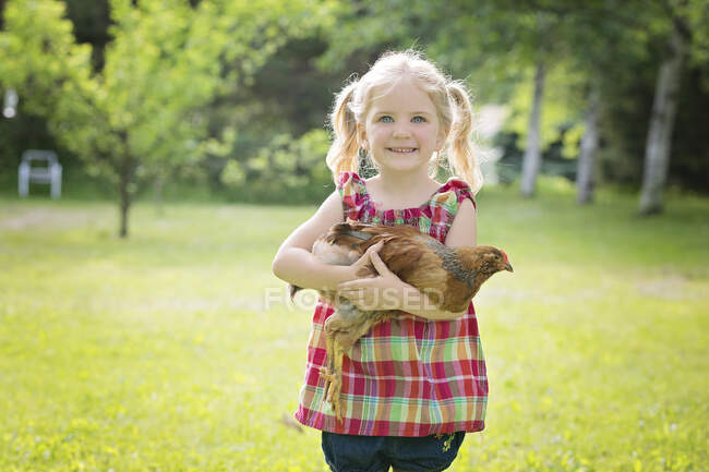 Cute little blond girl with blue eyes holding a chicken outdoors. — Stock Photo
