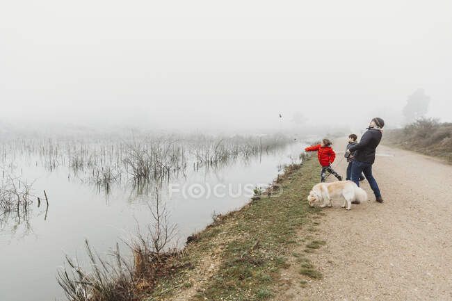 Pai e dois filhos com cão jogando pedras no lago no dia nebuloso — Fotografia de Stock