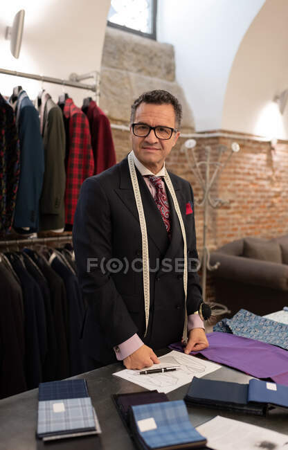 Mature male tailor standing near table with sketch and cloth samples in atelier — Stock Photo