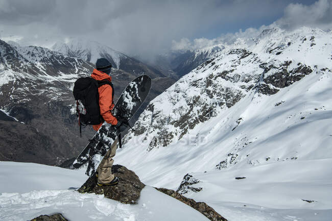 Homme avec snowboard debout sur la roche à la montagne enneigée contre ciel nuageux — Photo de stock