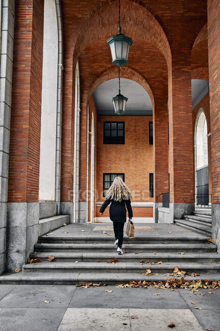 Vue arrière de la femelle blonde avec sac en papier marchant sur les marches dans l'arche du bâtiment en brique le jour de l'automne — Photo de stock