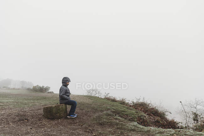 Garçon assis sur le poteau de béton en manteau d'hiver contre le ciel couvert brumeux — Photo de stock