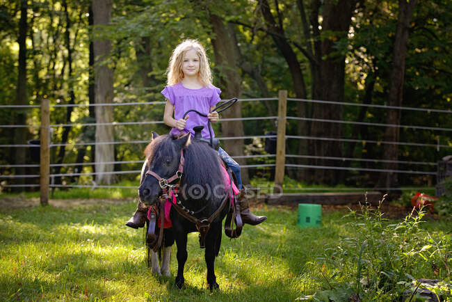 Schöne blonde Mädchen in lila Hemd reitet ein kleines schwarzes Pony. — Stockfoto