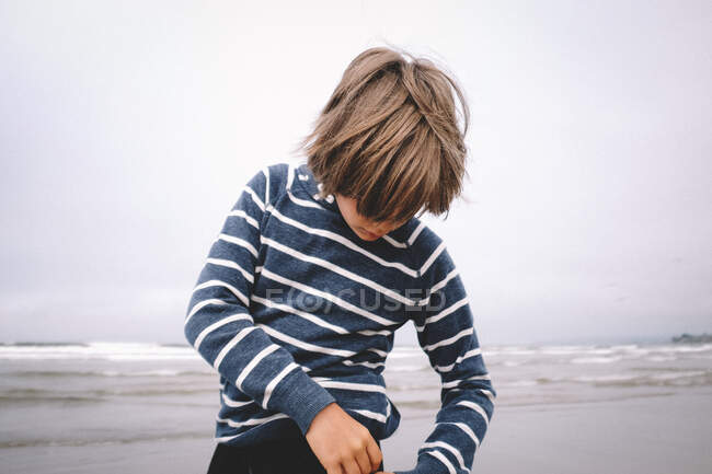 Menino de camisa listrada com cabelo comprido em uma praia — Fotografia de Stock