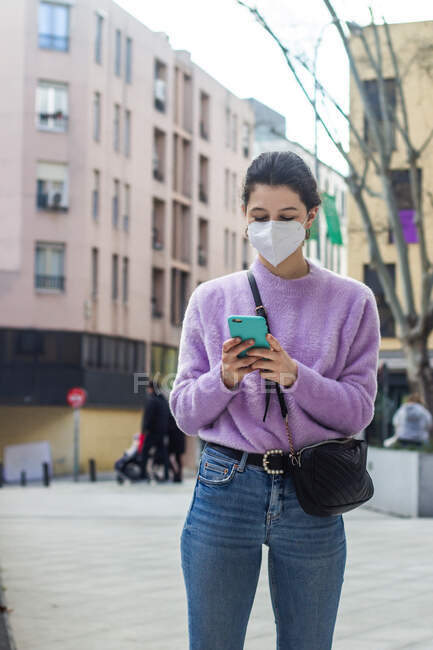 Молода жінка з захисною маскою KN95 на вулиці дивиться мобільний — стокове фото