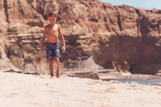 Niño en edad escolar caminando solo en una playa con acantilados en el fondo. - foto de stock