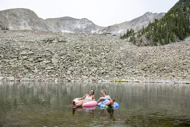 Felices amigas sentadas en anillos inflables en el lago durante las vacaciones - foto de stock