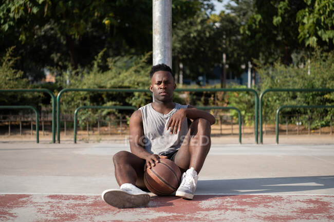 African American athlete with ball sitting near pole and resting on sports ground — Stock Photo