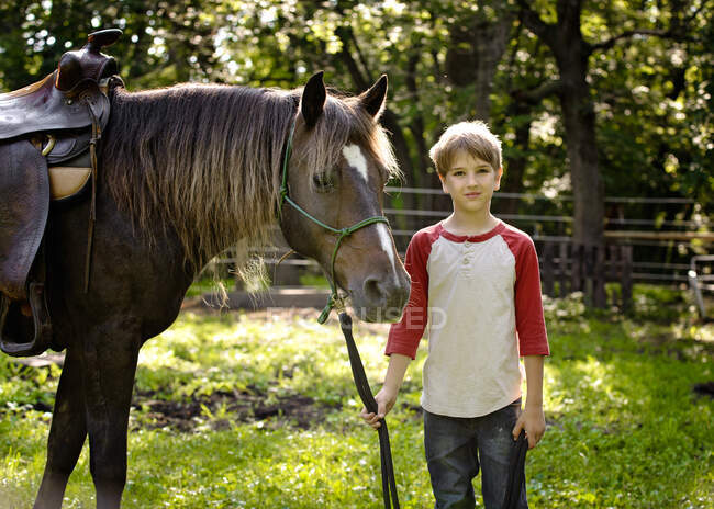 Handsome blond boy holding the reigns of horse in the country. — Stock Photo