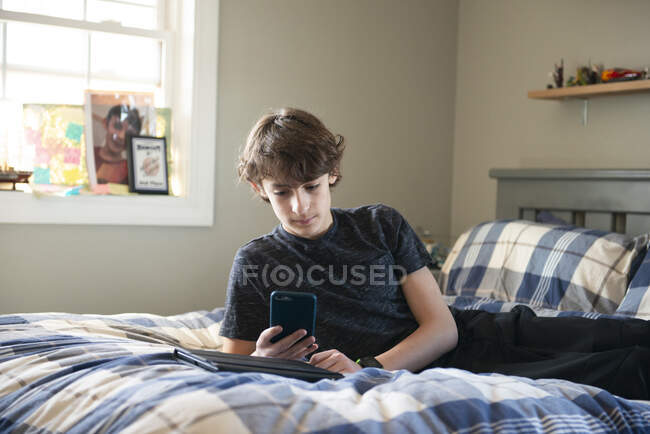 Tween menino deitado na cama com telefone e tablet. — Fotografia de Stock
