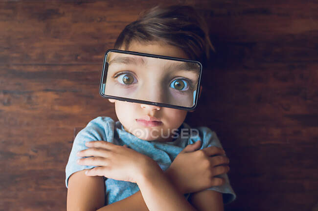 Boy laying on the ground with a phone photo of his eyes on top of face — Stock Photo
