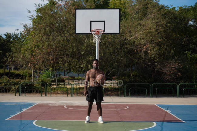Strong African American player with ball taking rest near basketball hoop on court — Fotografia de Stock