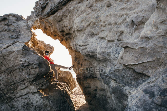Мальчик-подросток в маске для лица прислоняется к большой скале на пляже — стоковое фото