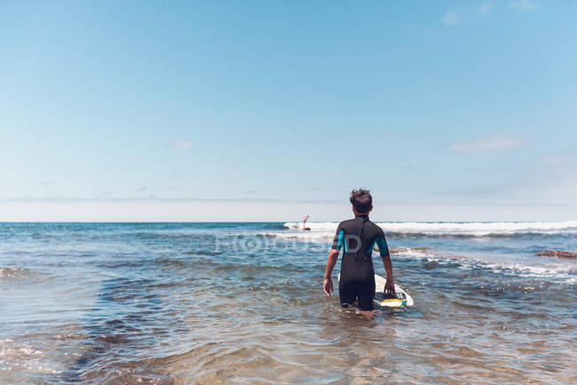 Young surfer entering the water while watching another surfer. — Stock Photo