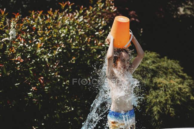 Boy standing pouring bucket of water over his own head — Stock Photo
