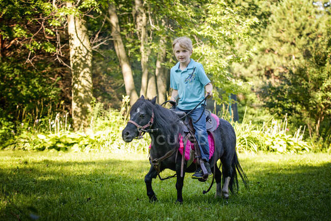 Piccolo biondo cavalcando il suo piccolo pony nero in campagna. — Foto stock