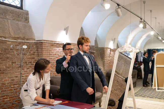 Mann passt Anzugjacke auf männliche Kundin in der Nähe von Spiegel und Assistentin macht Notizen — Stockfoto