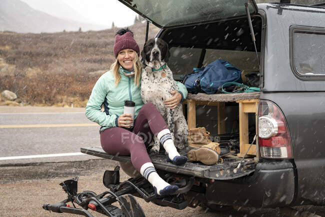 Smiling young woman sitting with dog in trunk of off-road vehicle during snowfall — Stock Photo