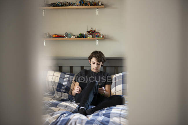 Ein Blick durch die Tür auf einen Jungen, der mit einem Tablet auf seinem Bett sitzt. — Stockfoto