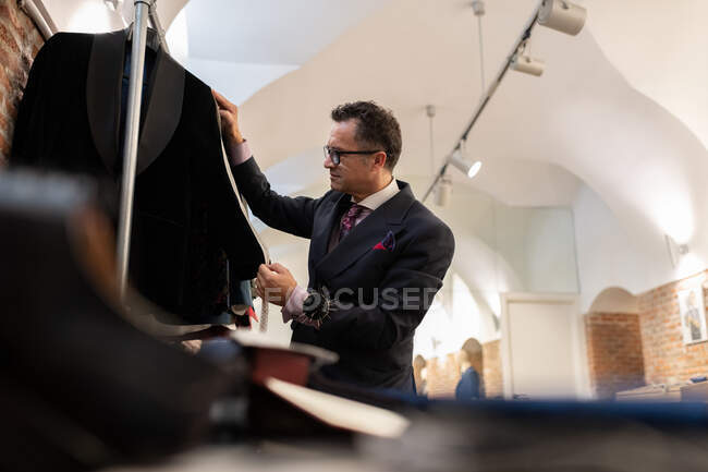 Manga de medição de alfaiate macho sênior de jaqueta elegante em rack durante o trabalho em estúdio — Fotografia de Stock