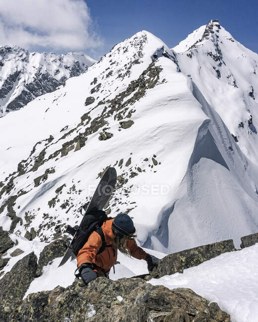 Hombre con snowboard escalando rocas en la montaña nevada durante las vacaciones - foto de stock