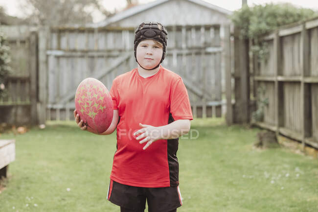 Junge trägt Rugby-Kopfbedeckung und hält Rugby-Ball im Hinterhof — Stockfoto