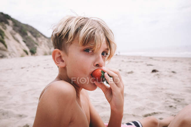 Blonder Junge isst eine Erdbeere an einem Sandstrand — Stockfoto