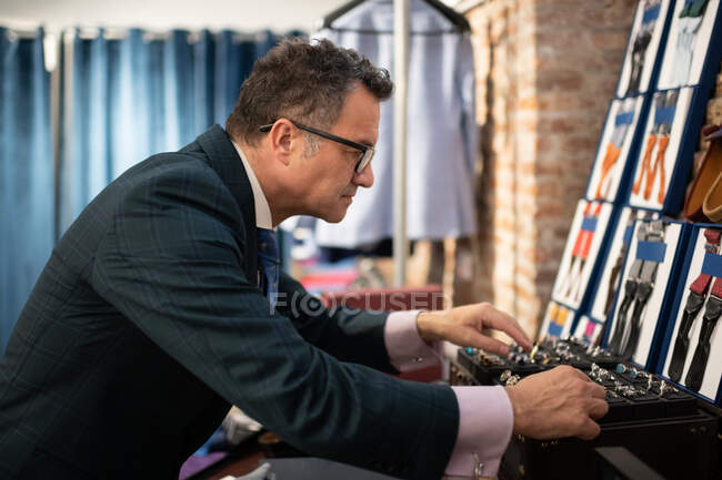Sarto uomo anziano in giacca e cravatta che seleziona accessori dalla scatola mentre lavora in atelier — Foto stock