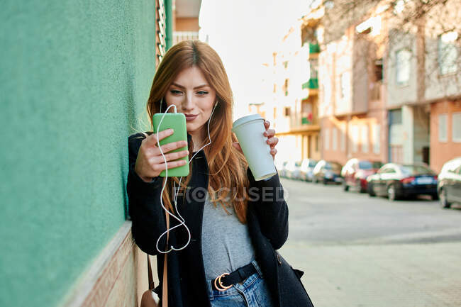 Mujer de negocios sosteniendo una taza de café mientras mira su teléfono escuchando música sobre un fondo verde - foto de stock