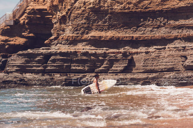Niño saliendo del agua llevando su tabla de surf - acantilados en la parte posterior. - foto de stock